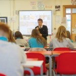 A teacher presenting to pupils in a classroom with red chairs and a large screen, surrounded by bilingual Welsh and English educational posters.