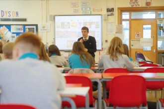 A teacher presenting to pupils in a classroom with red chairs and a large screen, surrounded by bilingual Welsh and English educational posters.