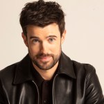Portrait of Jack Whitehall wearing a black jacket against a light background.