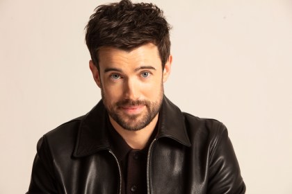 Portrait of Jack Whitehall wearing a black jacket against a light background.