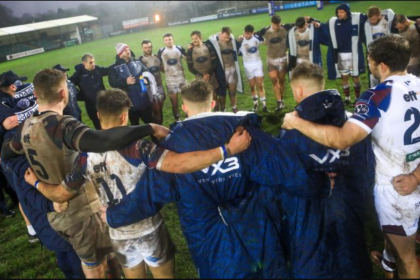 Swansea RFC players and coaches standing in a muddy post‑match huddle on the pitch at St Helen’s.