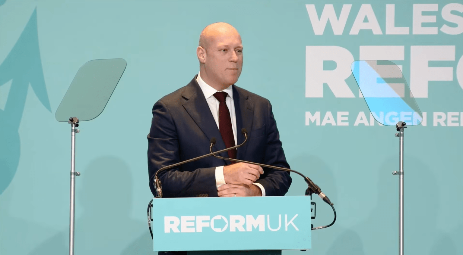 Dan Thomas standing at a Reform UK podium with Welsh branding behind him during a political rally.