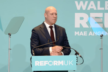 Dan Thomas standing at a Reform UK podium with Welsh branding behind him during a political rally.
