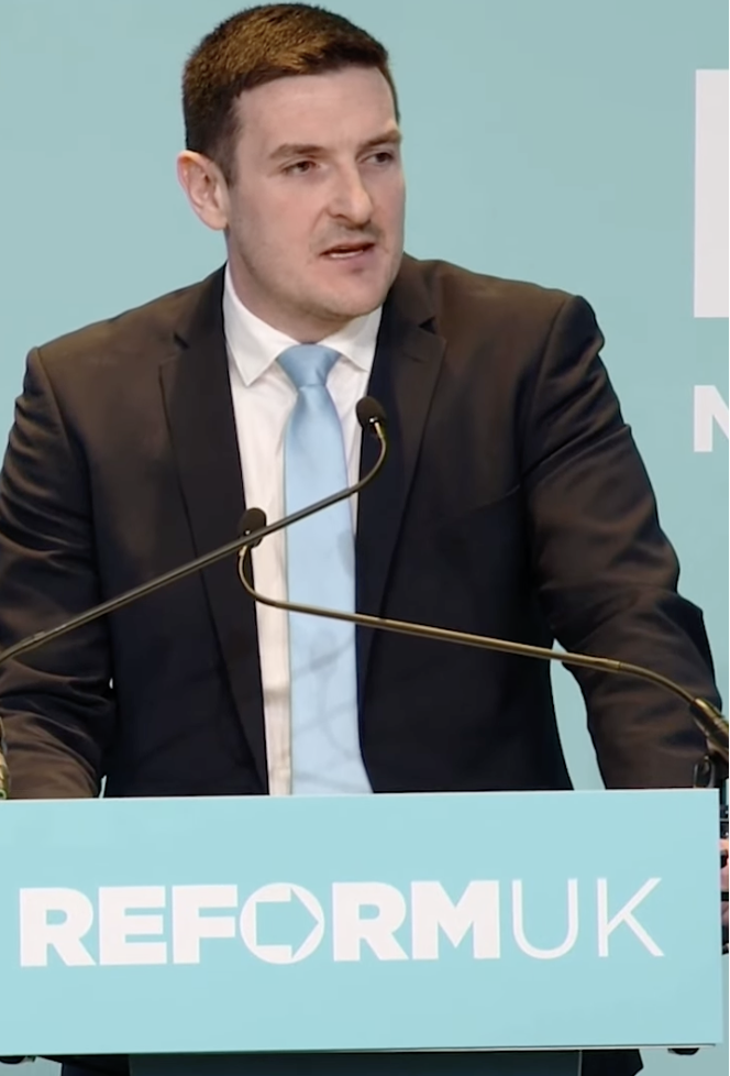 James Evans standing at a Reform UK podium during a speech following his defection from the Conservatives.