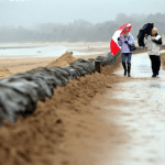 Two people walk along a rain‑soaked Swansea promenade under umbrellas, with sandbags lining the path and grey skies overhead.