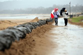 Two people walk along a rain‑soaked Swansea promenade under umbrellas, with sandbags lining the path and grey skies overhead.