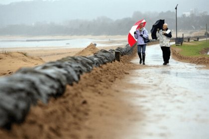 Two people walk along a rain‑soaked Swansea promenade under umbrellas, with sandbags lining the path and grey skies overhead.