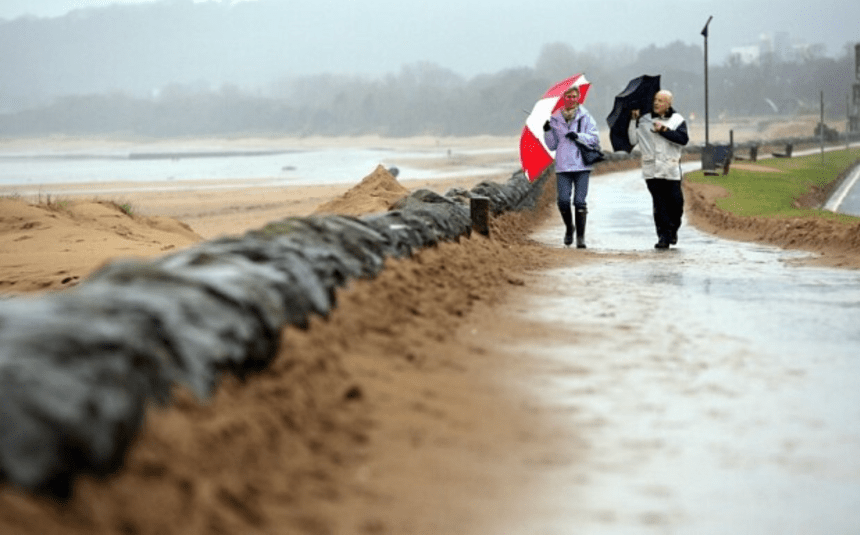 Two people walk along a rain‑soaked Swansea promenade under umbrellas, with sandbags lining the path and grey skies overhead.