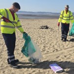 Two council workers in high‑visibility jackets collecting litter on Swansea Bay with large waste bags.