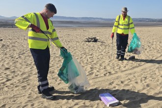 Two council workers in high‑visibility jackets collecting litter on Swansea Bay with large waste bags.