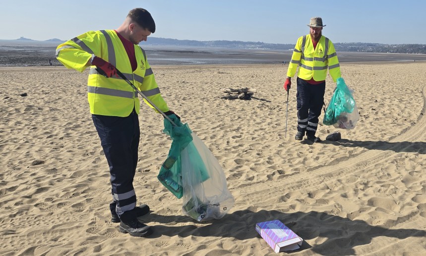 Two council workers in high‑visibility jackets collecting litter on Swansea Bay with large waste bags.