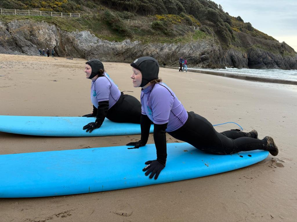 Two people in wetsuits and purple rash guards practicing surfing moves on blue boards on a sandy beach with cliffs and ocean in the background.
