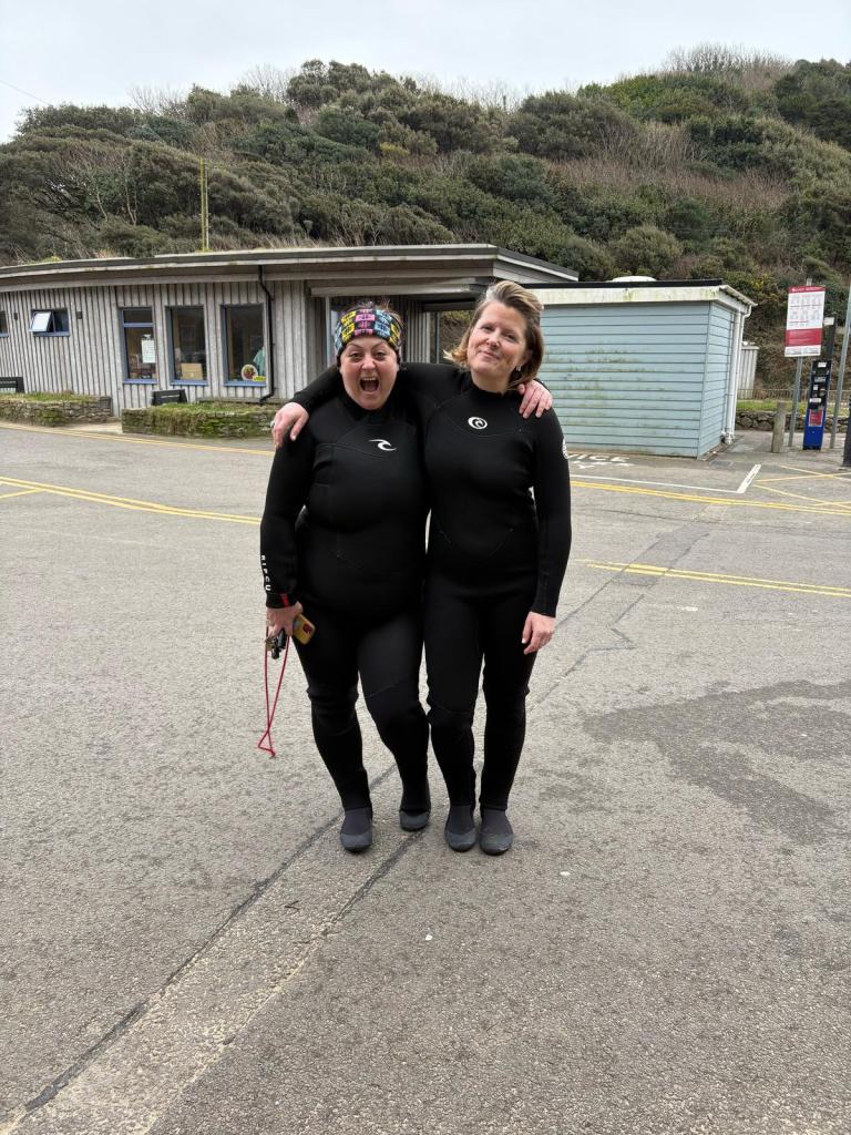 Two people in black wetsuits standing arm-in-arm near a wooden building and blue shed, with greenery behind them.