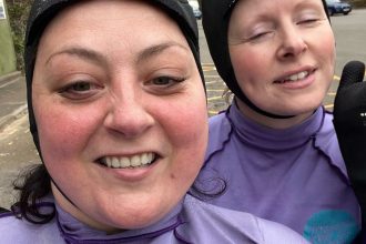 Two people in purple wetsuits and neoprene hoods smiling for a selfie in a car park with trees in the background.