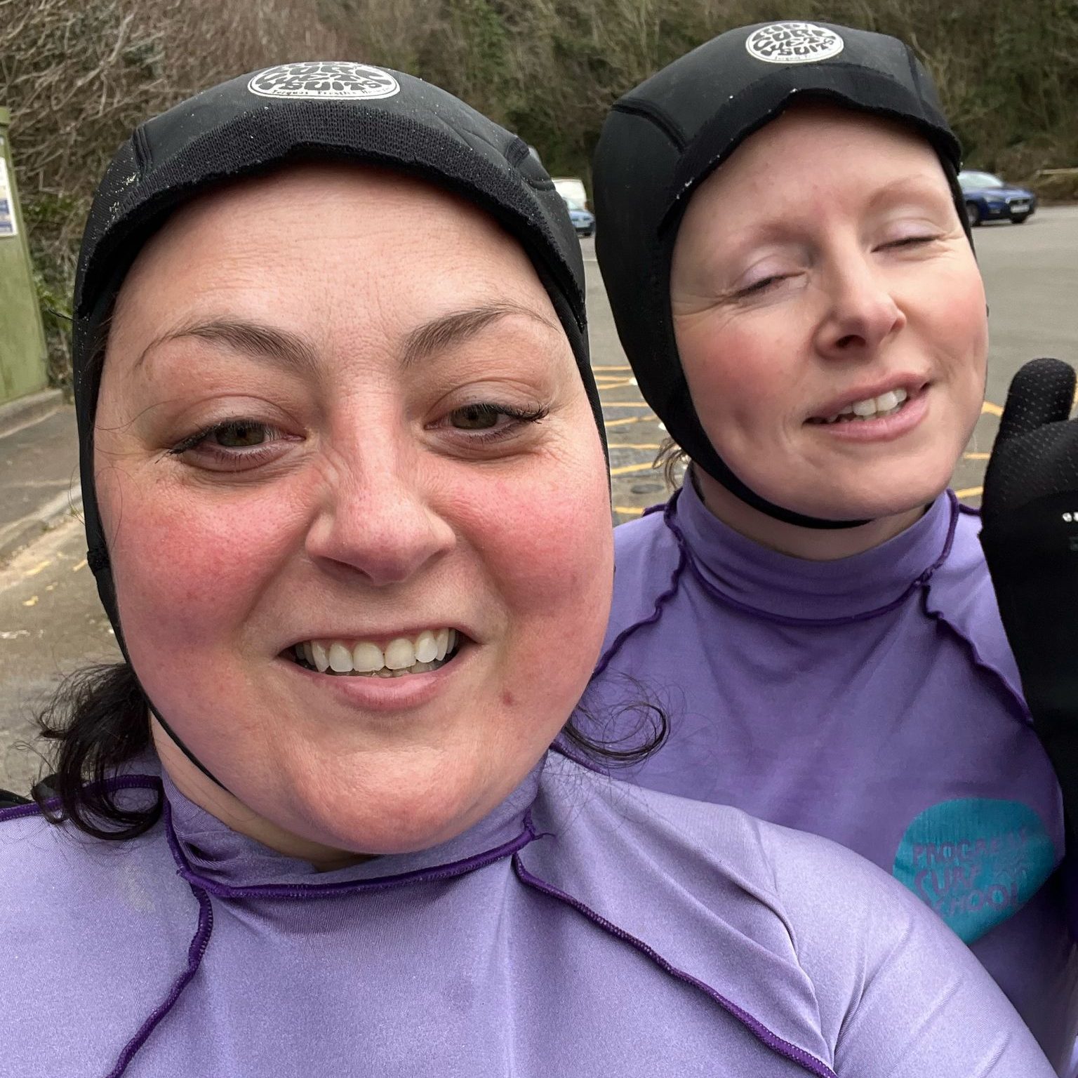 Two people in purple wetsuits and neoprene hoods smiling for a selfie in a car park with trees in the background.
