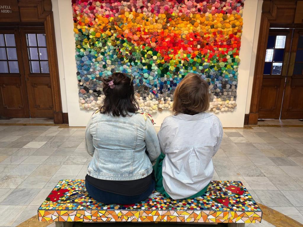 Two people seated on a mosaic bench facing a colourful wall art installation in a gallery space with wooden doors and marble flooring.
