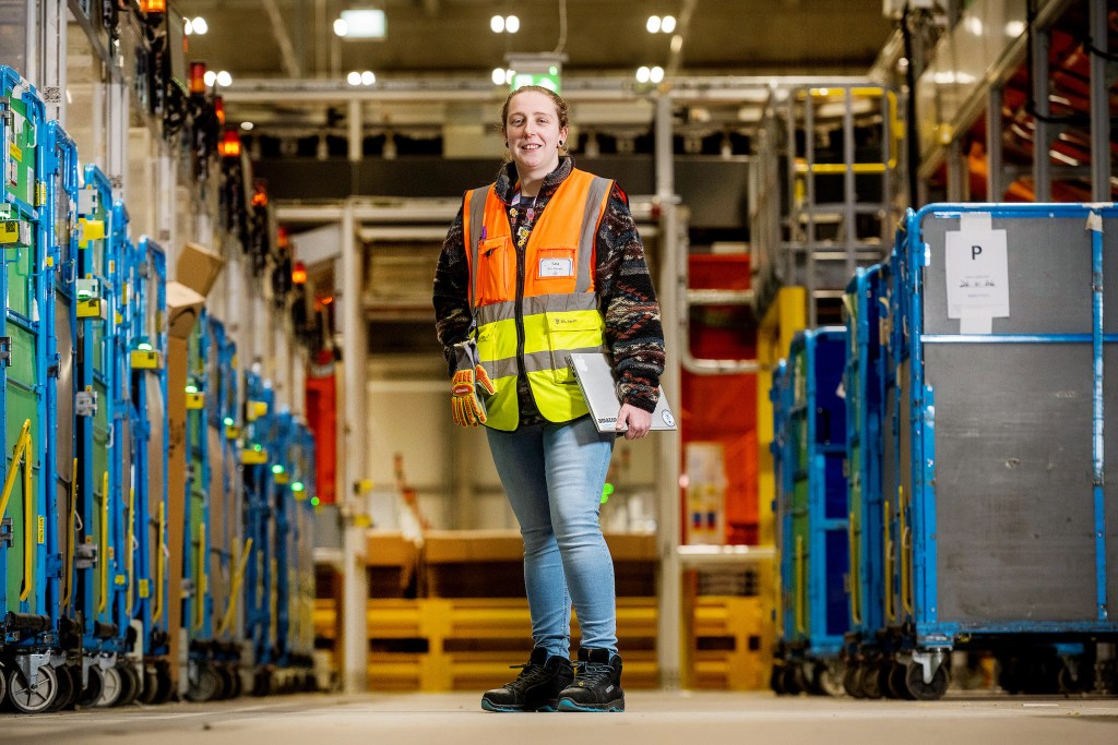 Sara Evans in a high‑visibility vest holding a clipboard inside Amazon’s Swansea fulfilment centre, surrounded by rows of rolling containers.