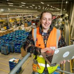 Sara Evans wearing a high‑visibility vest and holding a laptop on a platform inside Amazon’s Swansea fulfilment centre, overlooking rows of warehouse equipment.