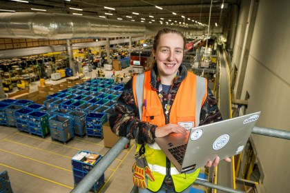 Sara Evans wearing a high‑visibility vest and holding a laptop on a platform inside Amazon’s Swansea fulfilment centre, overlooking rows of warehouse equipment.