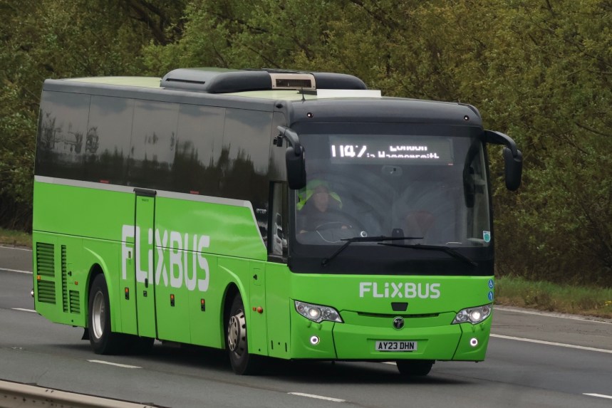 A green FlixBus coach travelling along a rural highway, displaying a digital destination sign for London.