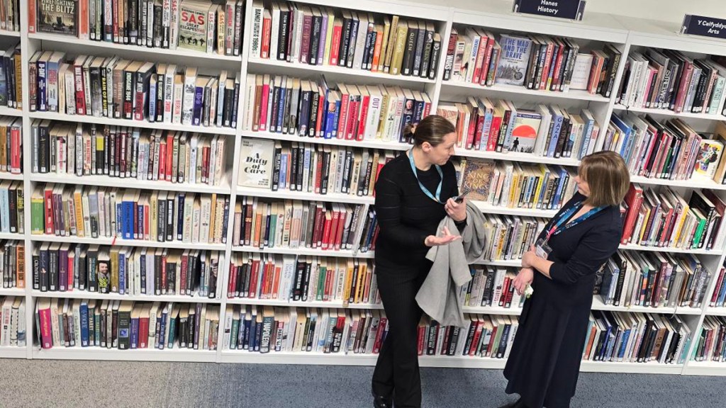 Two people talking in front of library shelves labelled “Hanes History” and “Y Celfyddydau Arts” at Y Storfa.