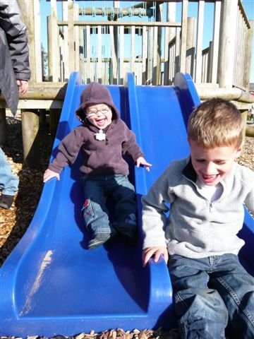 Two young brothers smiling as they go down a double slide at a playground.