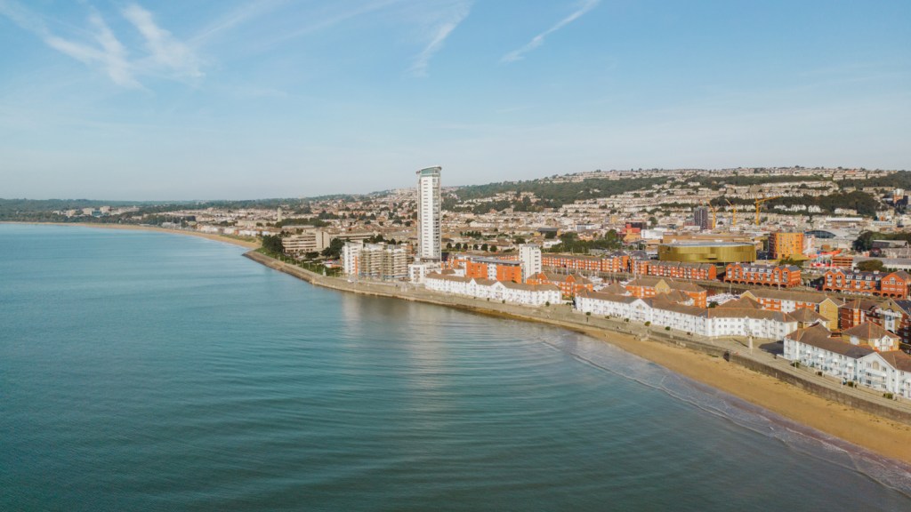 Coastal view of Swansea showing the beach, seafront buildings, the Meridian Tower and the city rising into the hills behind.