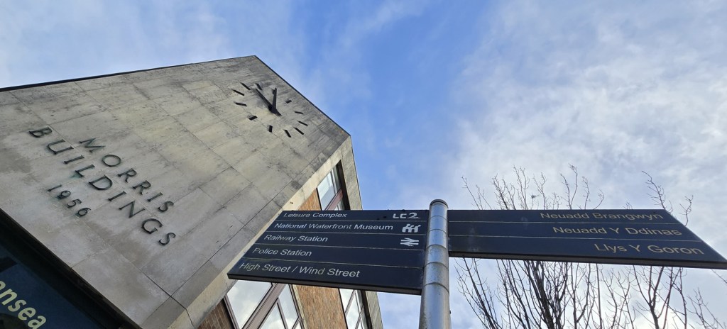 Morris Buildings in Swansea with a bilingual directional signpost showing routes to the LC, Waterfront Museum, railway station and other city locations.