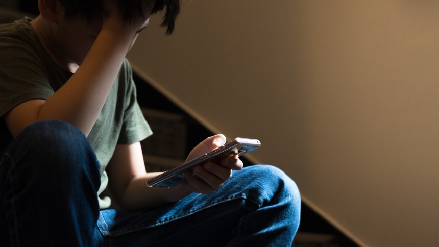 Teenager sitting on a staircase looking at a smartphone, appearing worried or overwhelmed, illustrating concerns about harmful online content.
