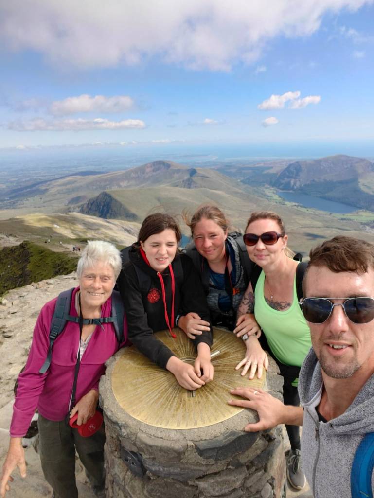 Group of five people standing at a mountain summit beside a topograph with panoramic views.