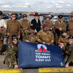 Group of people dressed in historical military uniforms from different eras standing with The Great Tommy Sleep Out banner at Swansea Airport