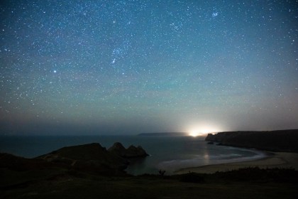 Star‑filled night sky above the Gower coastline with cliffs and shoreline visible.