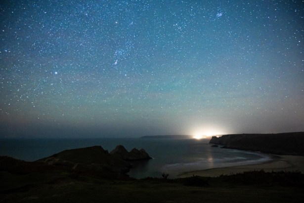 Star‑filled night sky above the Gower coastline with cliffs and shoreline visible.