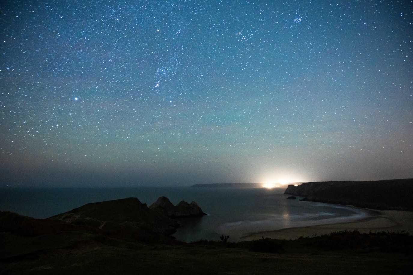Star‑filled night sky above the Gower coastline with cliffs and shoreline visible.