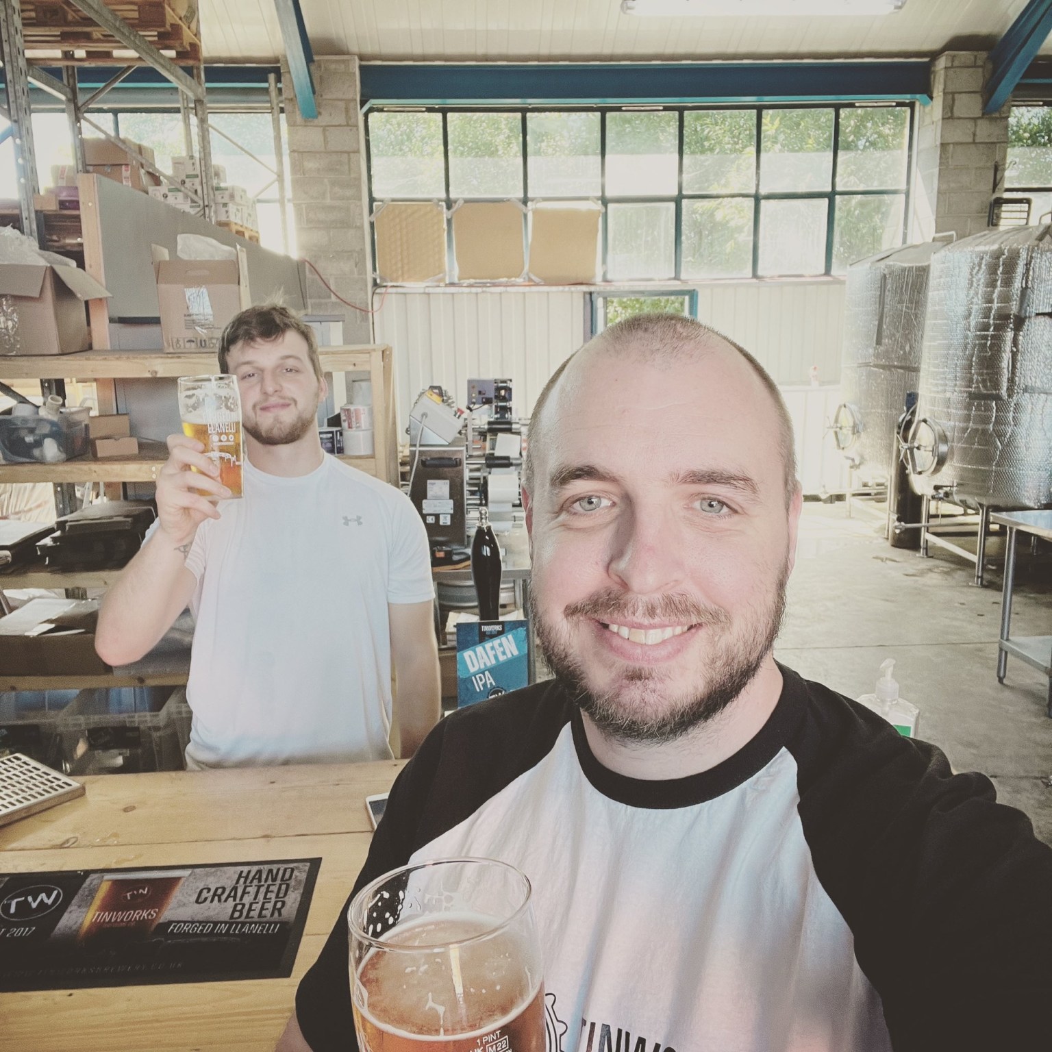 Two men smiling and holding pints of craft beer inside the Tinworks Brewery in Llanelli, with brewing tanks visible in the background