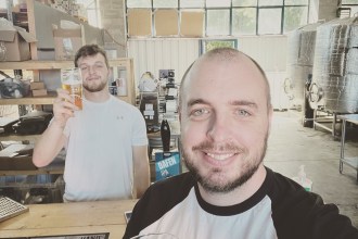 Two men smiling and holding pints of craft beer inside the Tinworks Brewery in Llanelli, with brewing tanks visible in the background