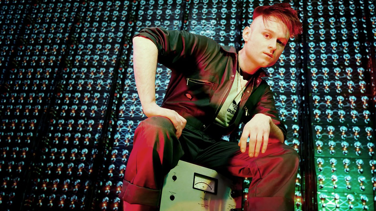 LOOK MUM NO COMPUTER sitting on vintage electronic equipment in a studio filled with illuminated components, photographed ahead of representing the UK at Eurovision 2026.