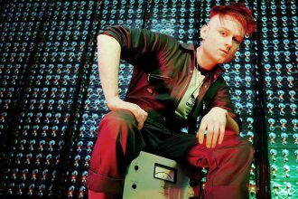 LOOK MUM NO COMPUTER sitting on vintage electronic equipment in a studio filled with illuminated components, photographed ahead of representing the UK at Eurovision 2026.