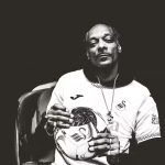 Cropped black‑and‑white image of Snoop Dogg wearing a Swansea City shirt and holding a football with the club badge.