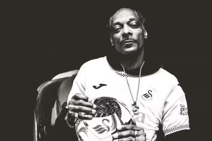 Cropped black‑and‑white image of Snoop Dogg wearing a Swansea City shirt and holding a football with the club badge.