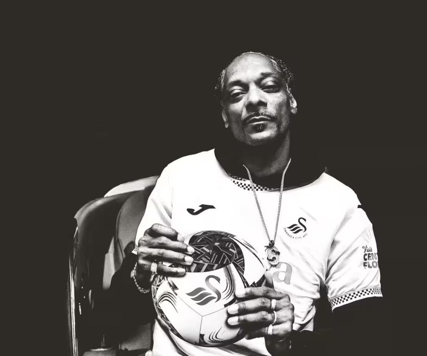Cropped black‑and‑white image of Snoop Dogg wearing a Swansea City shirt and holding a football with the club badge.