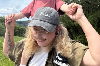 A smiling woman wearing glasses and a cap carries a young child on her shoulders outdoors, holding their hands as they play together