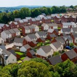 An elevated, wide-angle aerial shot of a large, modern housing estate with numerous red-brick and white-rendered houses, surrounded by lush green woodland under a clear sky.