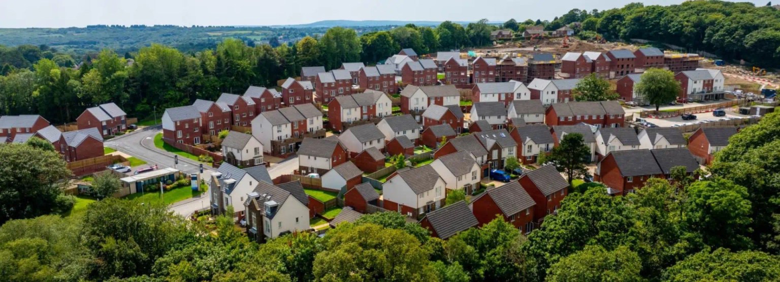 An elevated, wide-angle aerial shot of a large, modern housing estate with numerous red-brick and white-rendered houses, surrounded by lush green woodland under a clear sky.