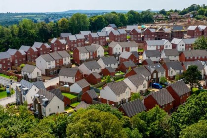 An elevated, wide-angle aerial shot of a large, modern housing estate with numerous red-brick and white-rendered houses, surrounded by lush green woodland under a clear sky.