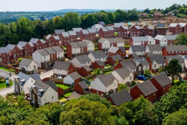 An elevated, wide-angle aerial shot of a large, modern housing estate with numerous red-brick and white-rendered houses, surrounded by lush green woodland under a clear sky.