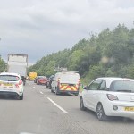 A photograph taken from inside a stationary vehicle on a dual carriageway, showing a queue of cars, vans and a lorry backed up ahead. A yellow recovery vehicle is visible further up the road. Dense green trees line both sides of the carriageway under an overcast sky.