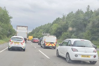 A photograph taken from inside a stationary vehicle on a dual carriageway, showing a queue of cars, vans and a lorry backed up ahead. A yellow recovery vehicle is visible further up the road. Dense green trees line both sides of the carriageway under an overcast sky.