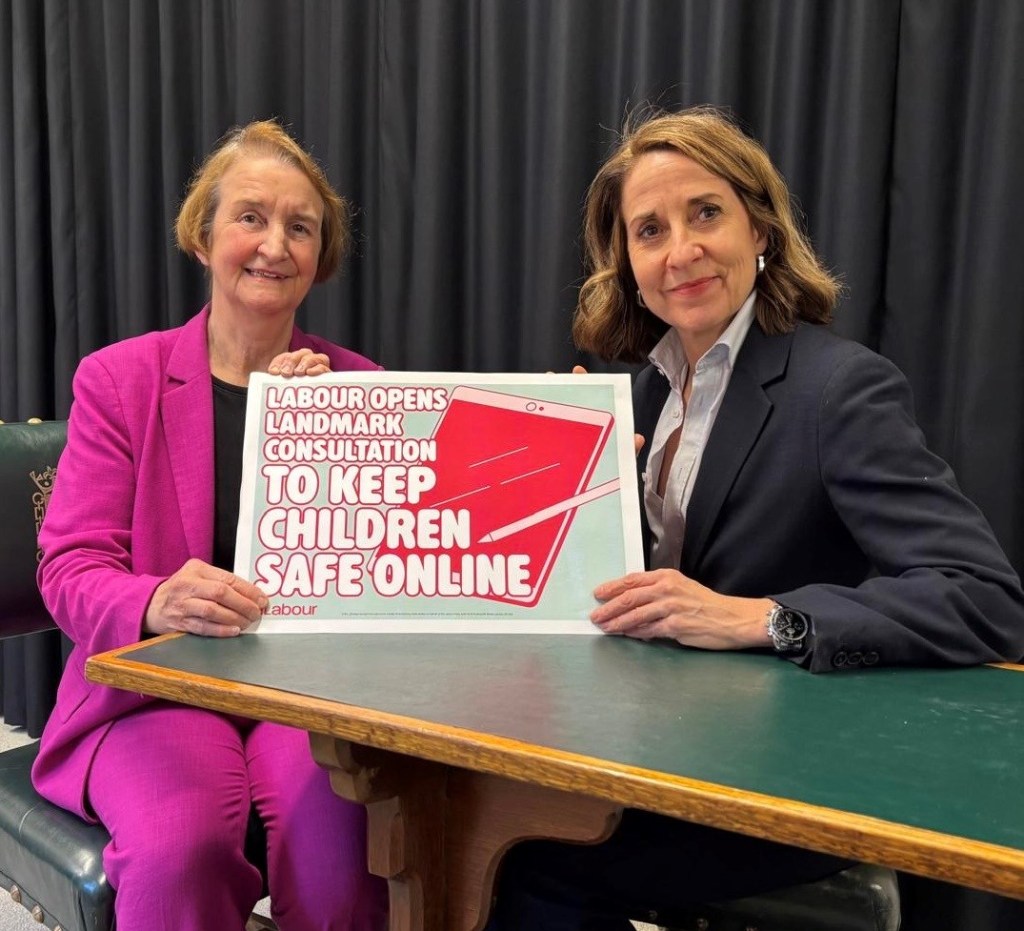 Dame Nia Griffith MP, in a bright pink trouser suit, and Technology Secretary Liz Kendall, in a dark suit, sit at a table holding a sign that reads "Labour opens landmark consultation to keep children safe online".