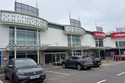 The exterior of Parc Fforestfach retail park showing the vacant units 4 and 5 in the foreground, with The Food Warehouse and a Jollyes "coming soon" sign visible to the right, and cars parked in the foreground.